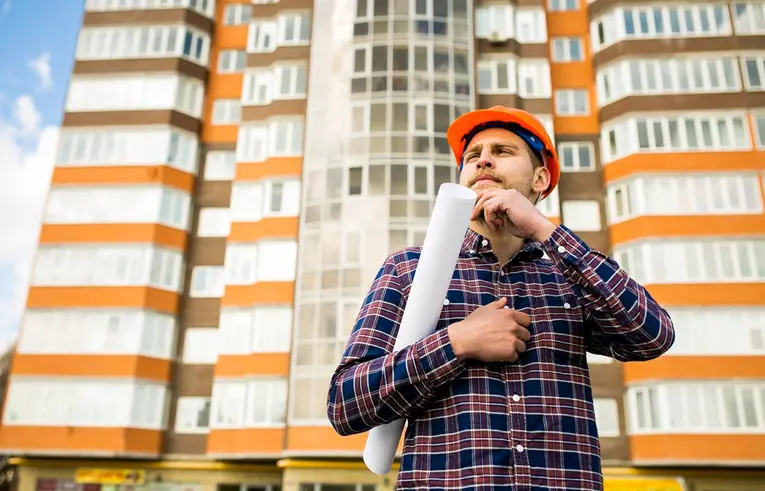 Mężczyzna we fioletowej flanelowej koszuli i czerwonym kasku budowlanym przed blokiem mieszkalnym, pod pachą trzyma zwinięty arkusz papieru
