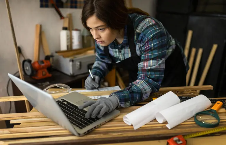 Kobieta w warsztacie stolarskim, ubrana w odzież roboczą, patrzy na ekran laptopa
