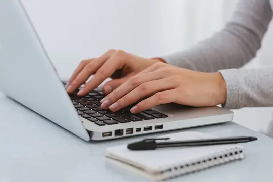 Dłonie na laptopie, obok leży notes i długopis
