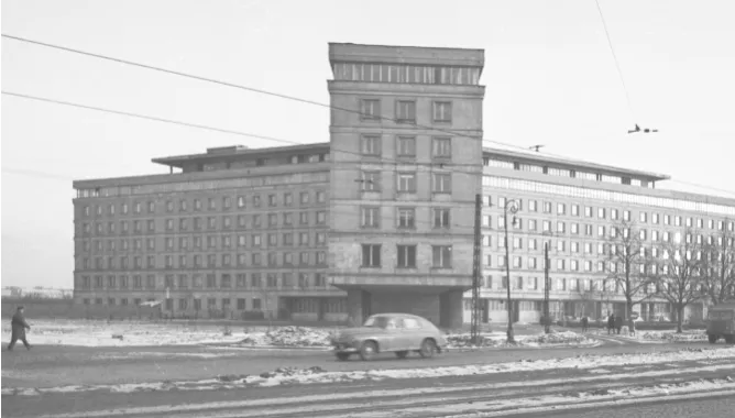 Czarno-białe zdjęcie budynku Głównego Urzędu Statystycznego w Warszawie. Na tle Urzędu widoczny jest samochód osobowy. 